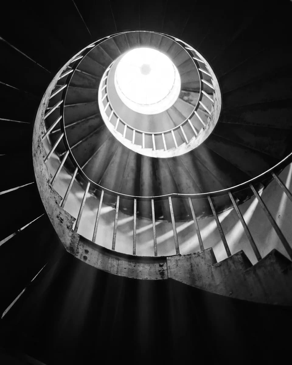 A spiral staircase seen from below, winding up toward a bright circular skylight