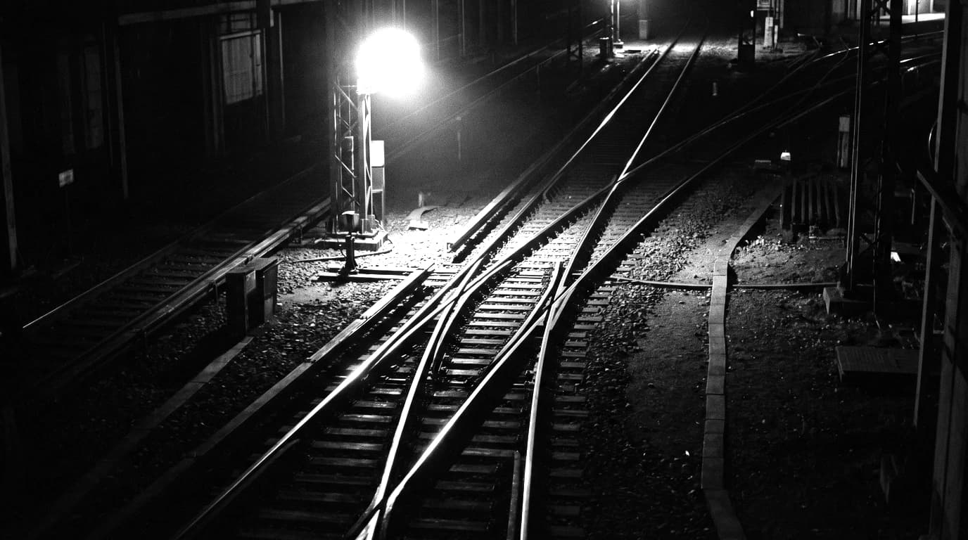 Black-and-white still of a railway junction at night, one rail splitting into three tracks under a single bright lamp